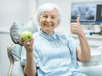 Senior woman holding apple and giving thumbs up in dental chair